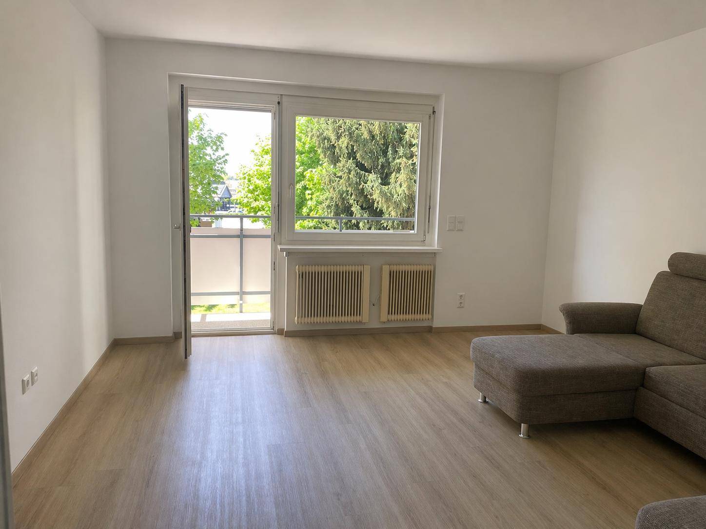 Geräumiges Wohnzimmer mit Laminatboden, Sofa und Zugang zum Balkon.