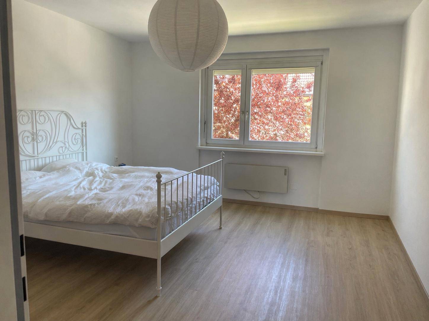 Schlafzimmer mit weißem Metallbett, Laminatboden und Blick ins Grüne.