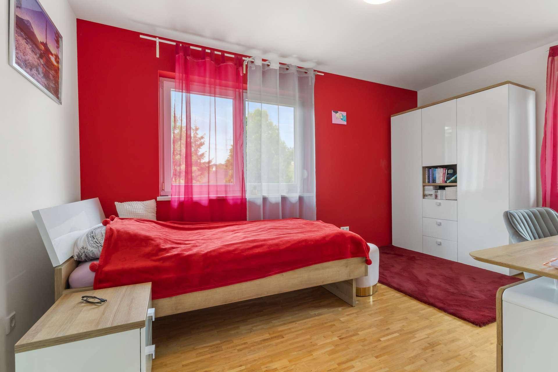 Farbenfrohes Schlafzimmer mit roter Akzentwand, weißem Kleiderschrank und Fenster.