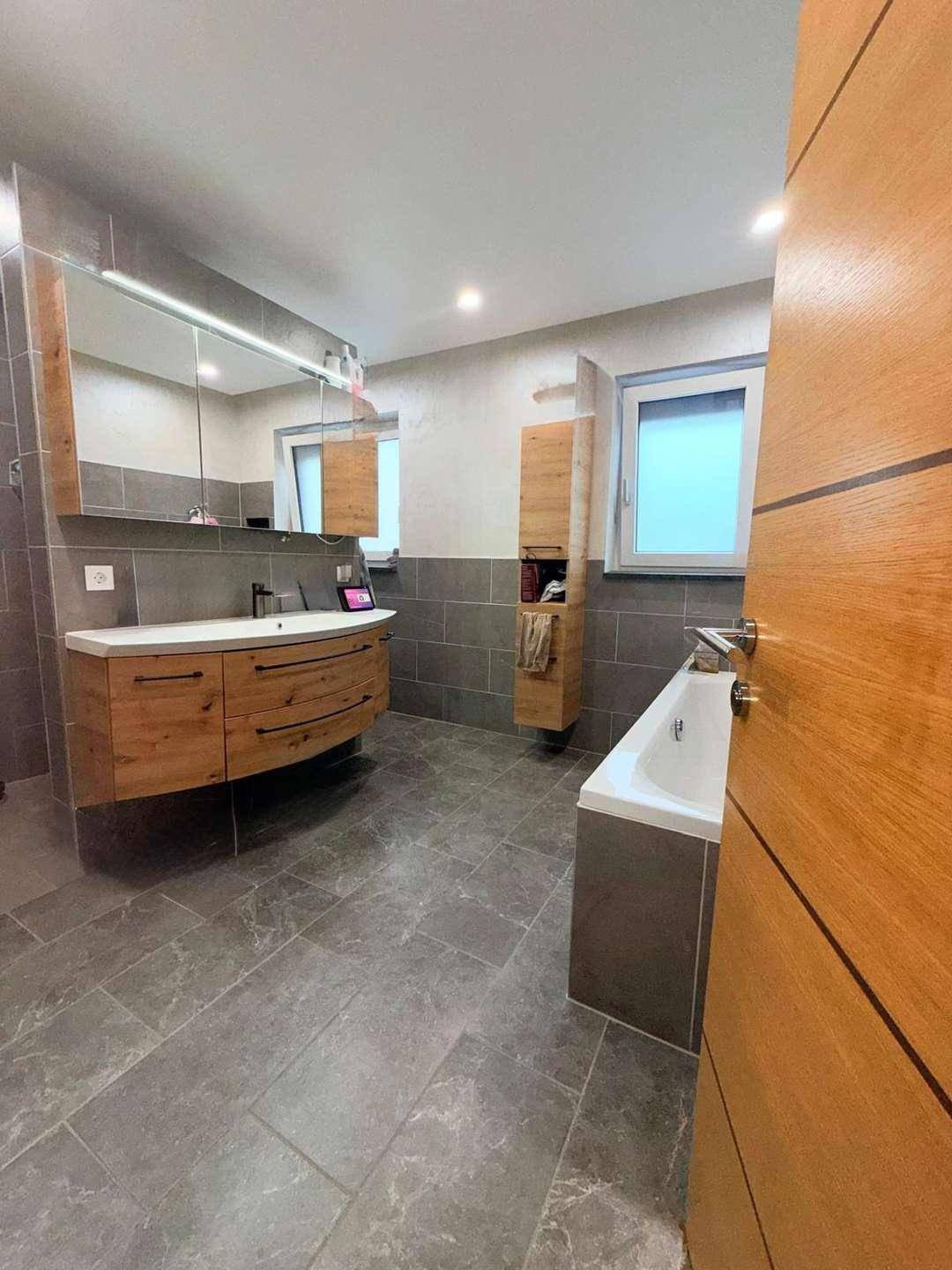 Modernes Badezimmer mit Holzwaschtisch, großer Badewanne und grauen Fliesen.