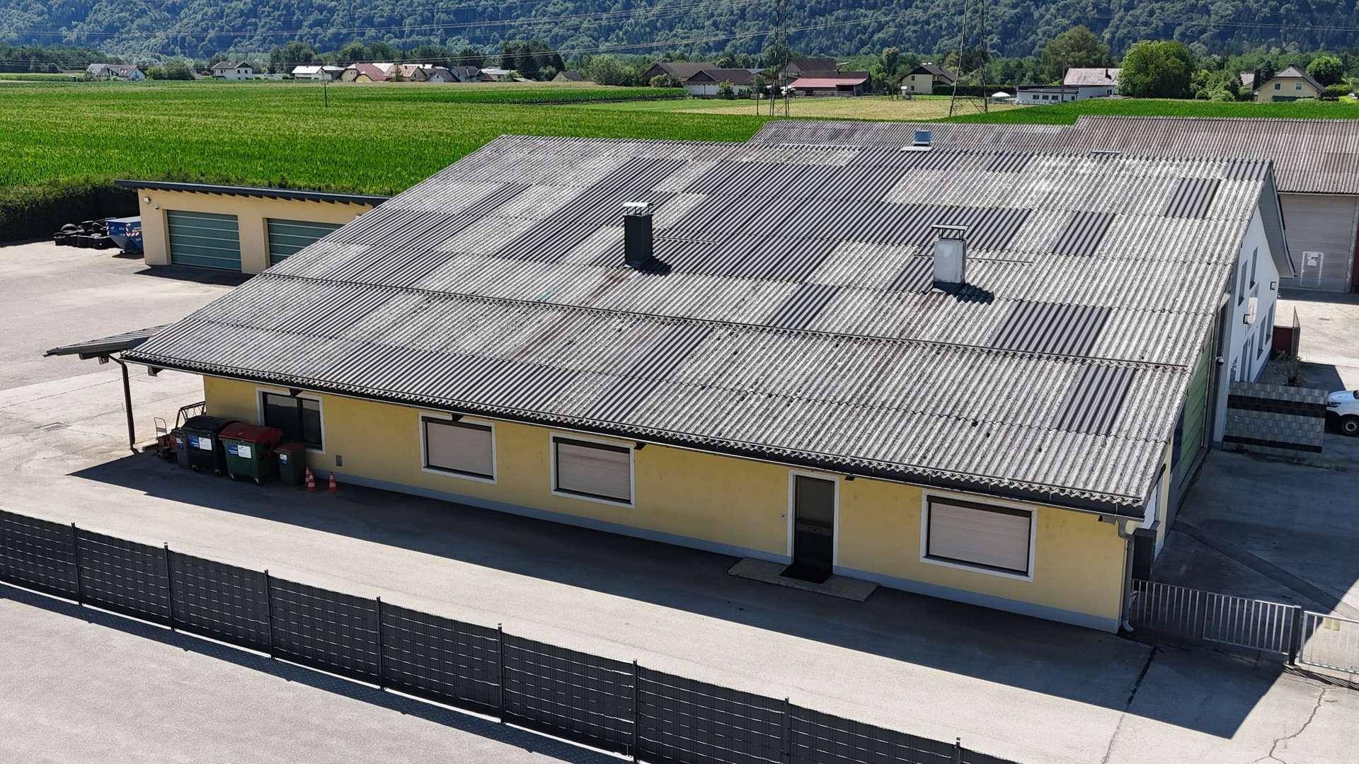 Langes, gelbes Gebäude mit Wellblechdach und großem Vorplatz, umgeben von Feldern und Bergen.