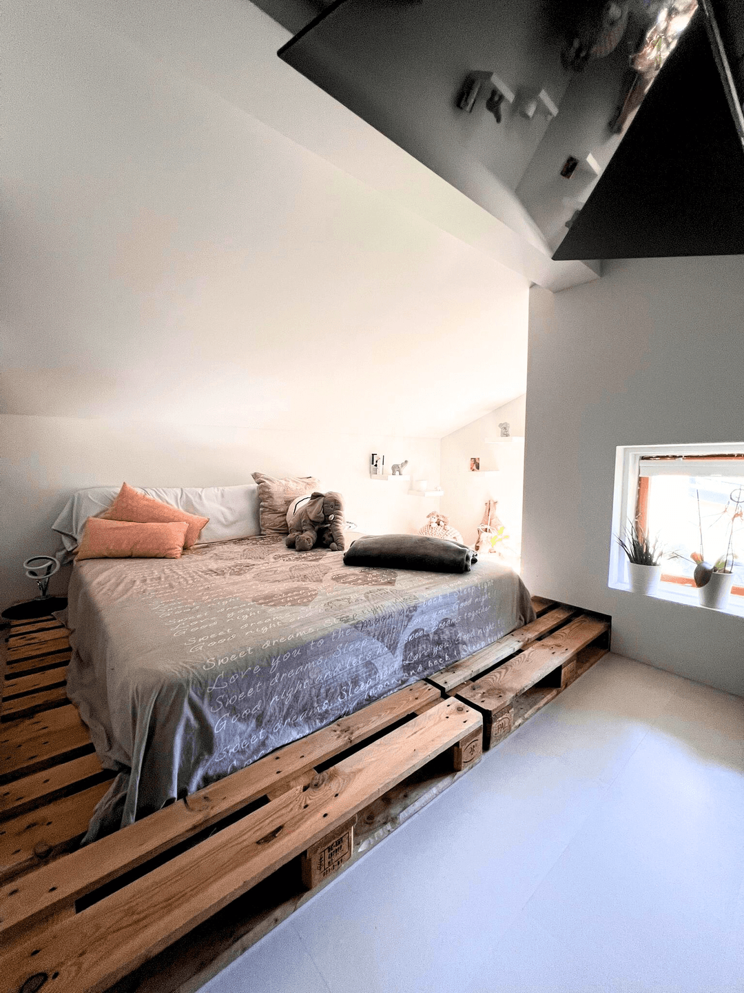 Schlafbereich im Dachgeschoss mit Palettenbett, kleinem Fenster und schrägen Wänden.