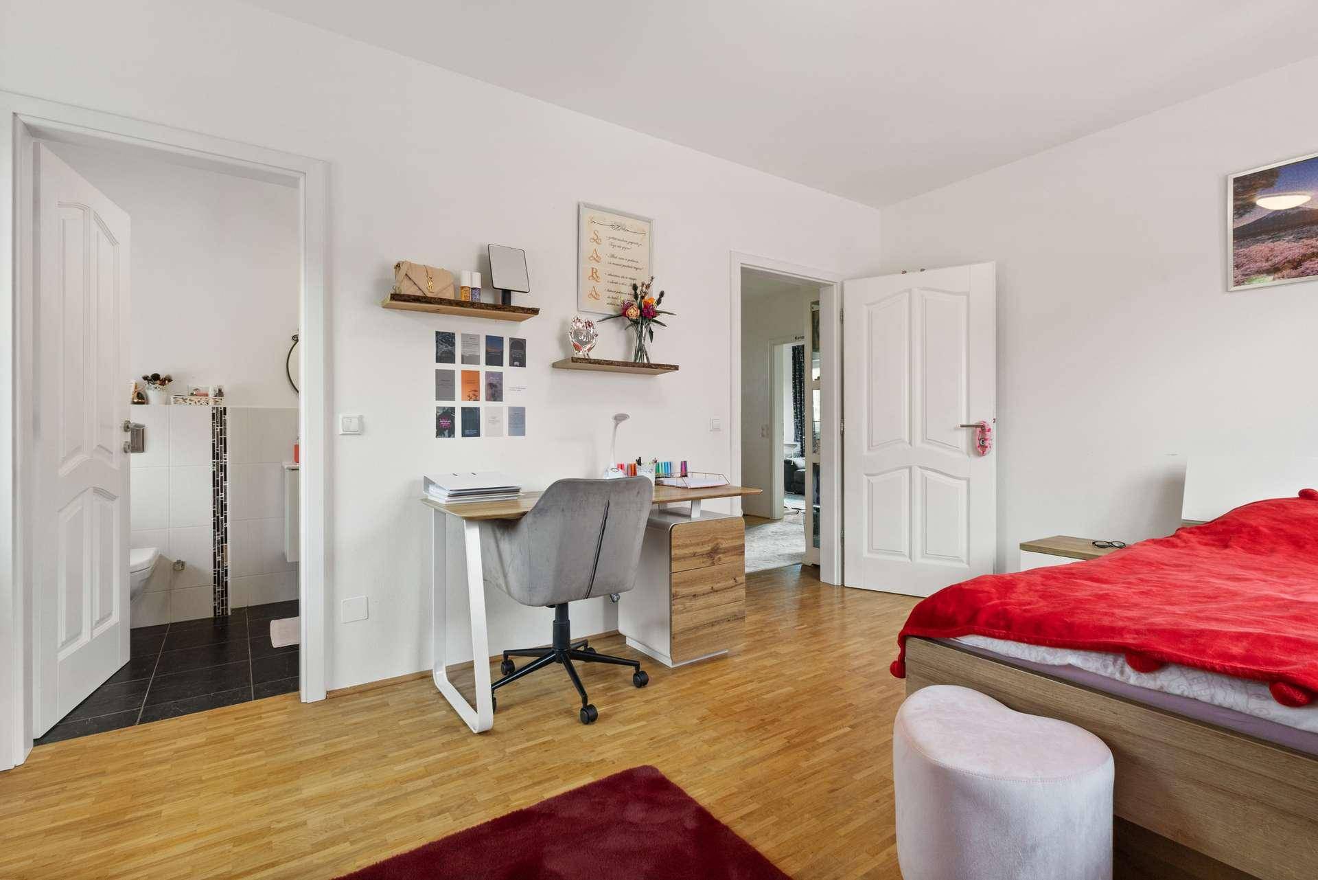 Helles Schlafzimmer mit rotem Bett, Schreibtisch und direktem Zugang zum Badezimmer.
