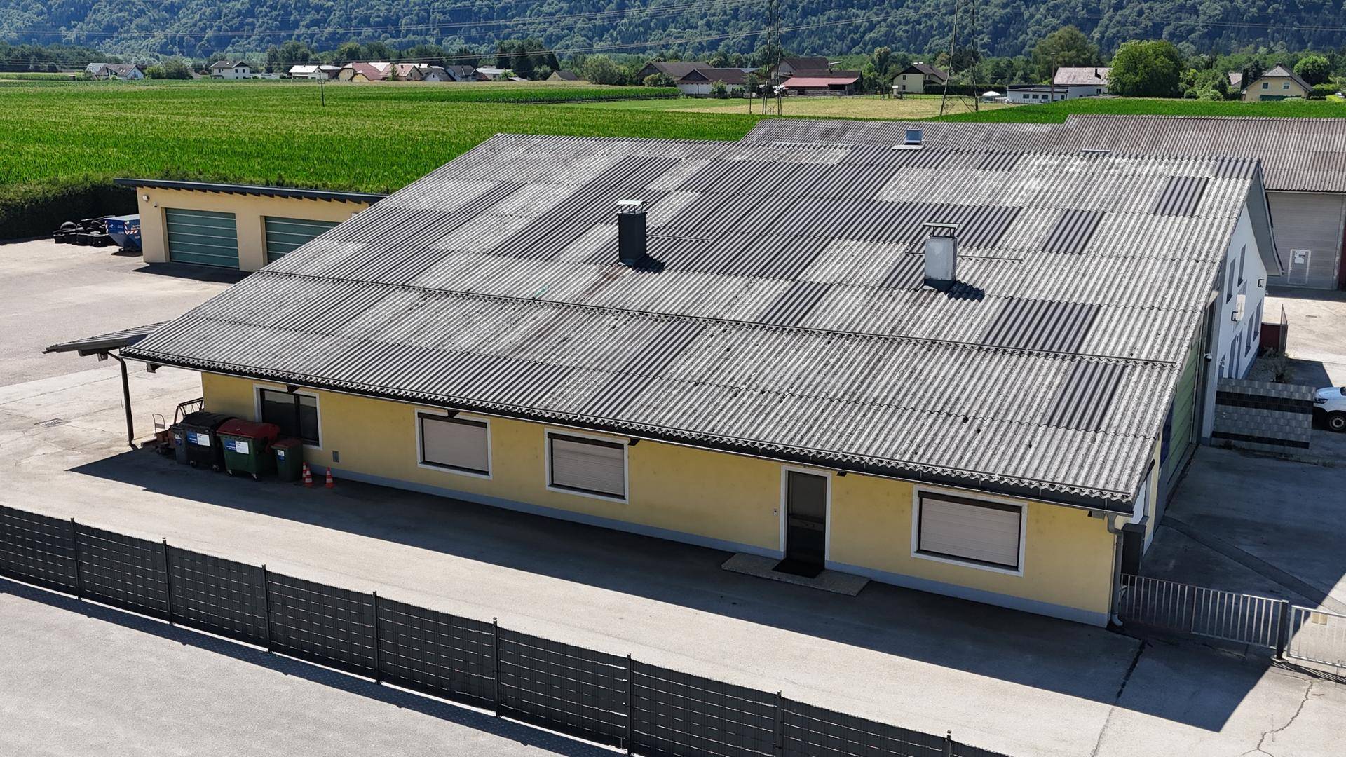 Luftaufnahme eines Gewerbegebäudes mit großem Wellblechdach und angrenzendem Parkplatzbereich in ländlicher Umgebung.