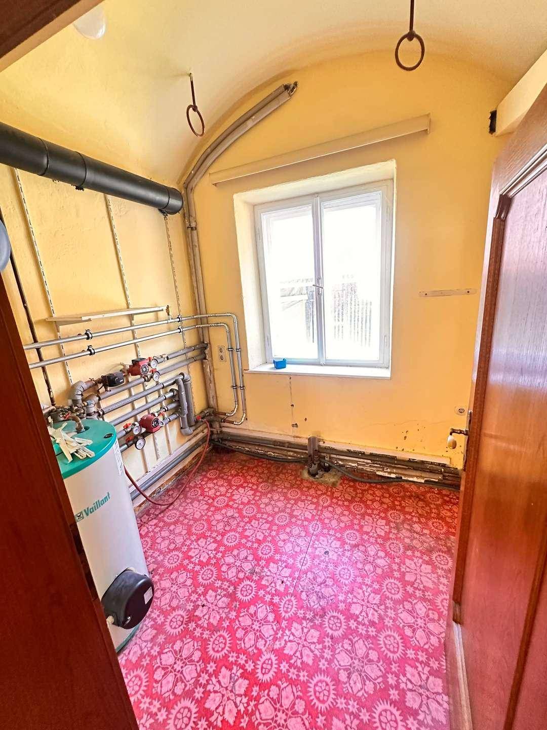 Hauswirtschaftsraum mit Warmwasserbereiter, sichtbaren Rohren und einem Fenster, roter Linoleumboden.