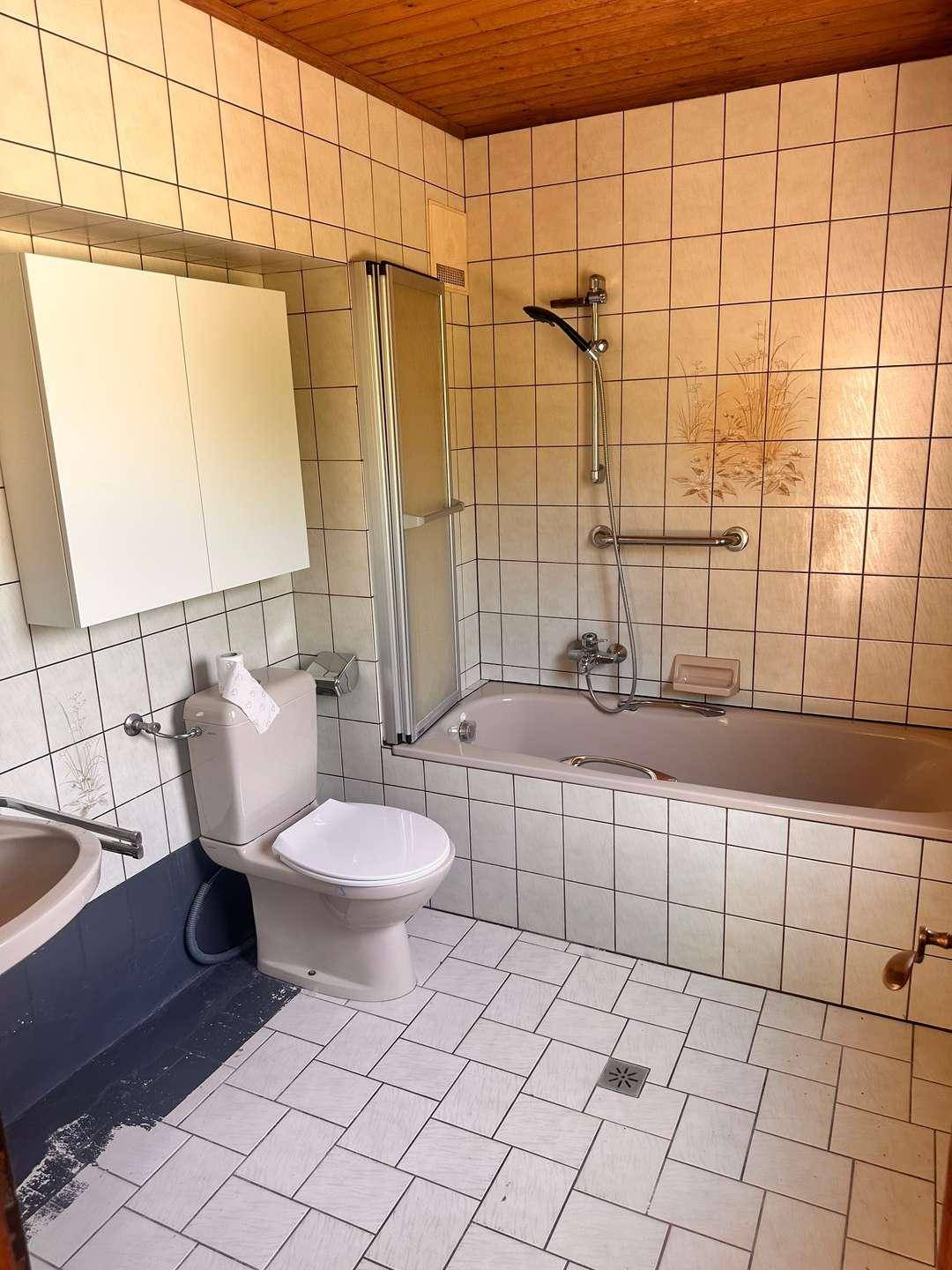 Badezimmer mit weißen Fliesen, Badewanne mit Duschfunktion und WC, ausgestattet mit Haltegriffen.