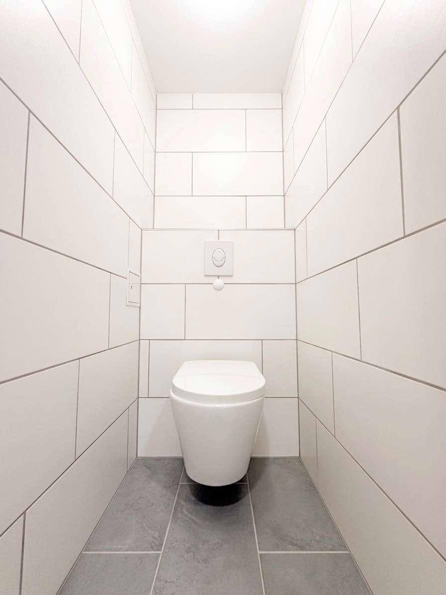 Separates WC mit modernen weißen Wandfliesen und grauen Bodenfliesen.