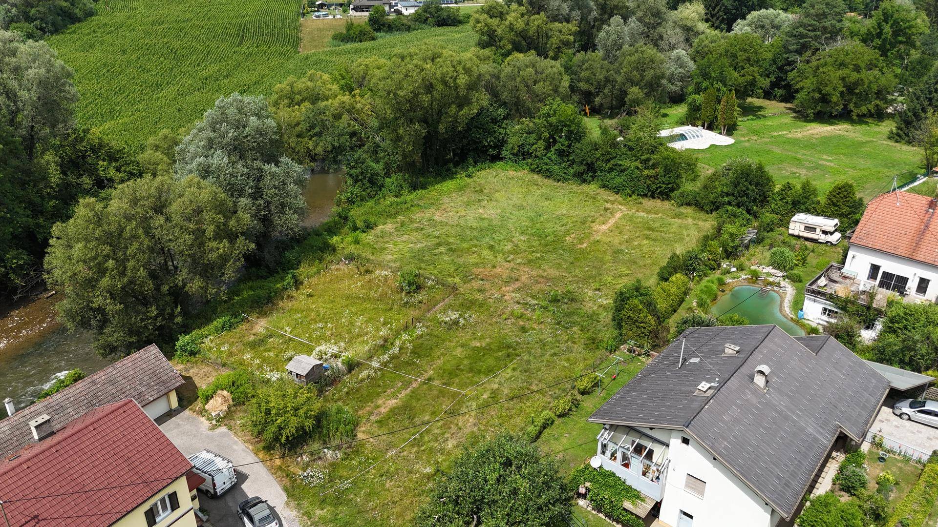 Vogelperspektive auf ein Grundstück mit angrenzendem Fluss, umgeben von grüner Landschaft und Häusern.