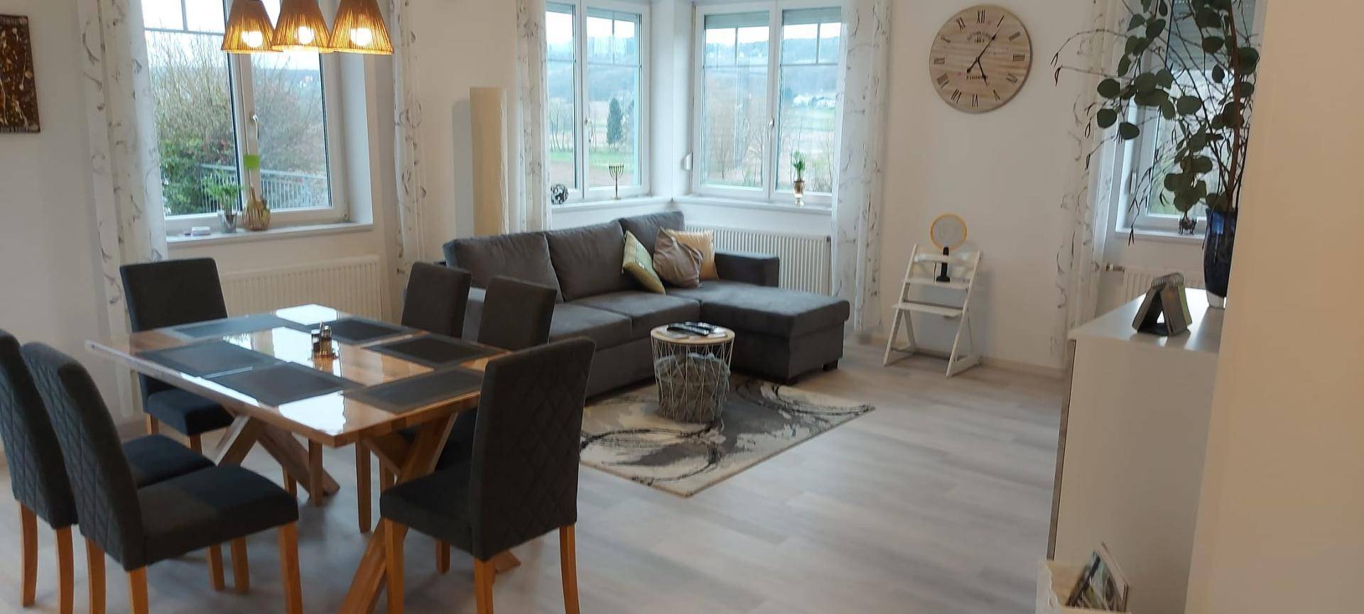 Heller Wohn- und Essbereich mit großen Fenstern, modernem Sofa und einem Holztisch mit dunklen Stühlen.