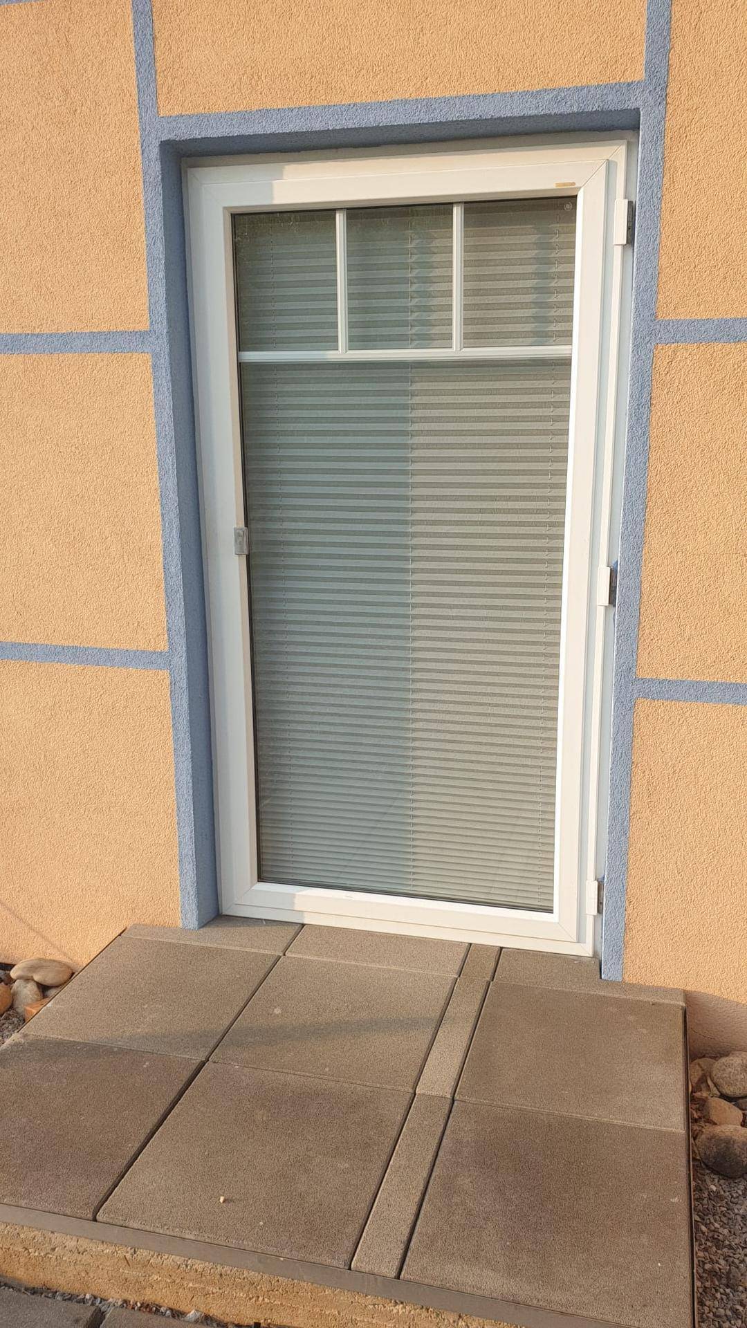 Nahaufnahme einer modernen weißen Tür mit Jalousien und einem kleinen gepflasterten Eingangsbereich.