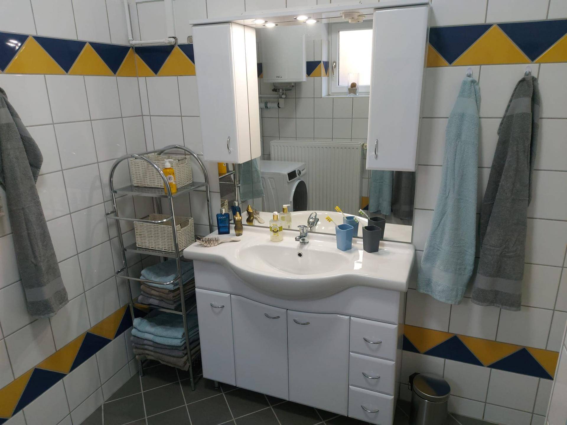 Badezimmer mit Waschtisch, Spiegel und farbenfrohen Fliesen in Gelb und Blau, sowie einem Fenster.
