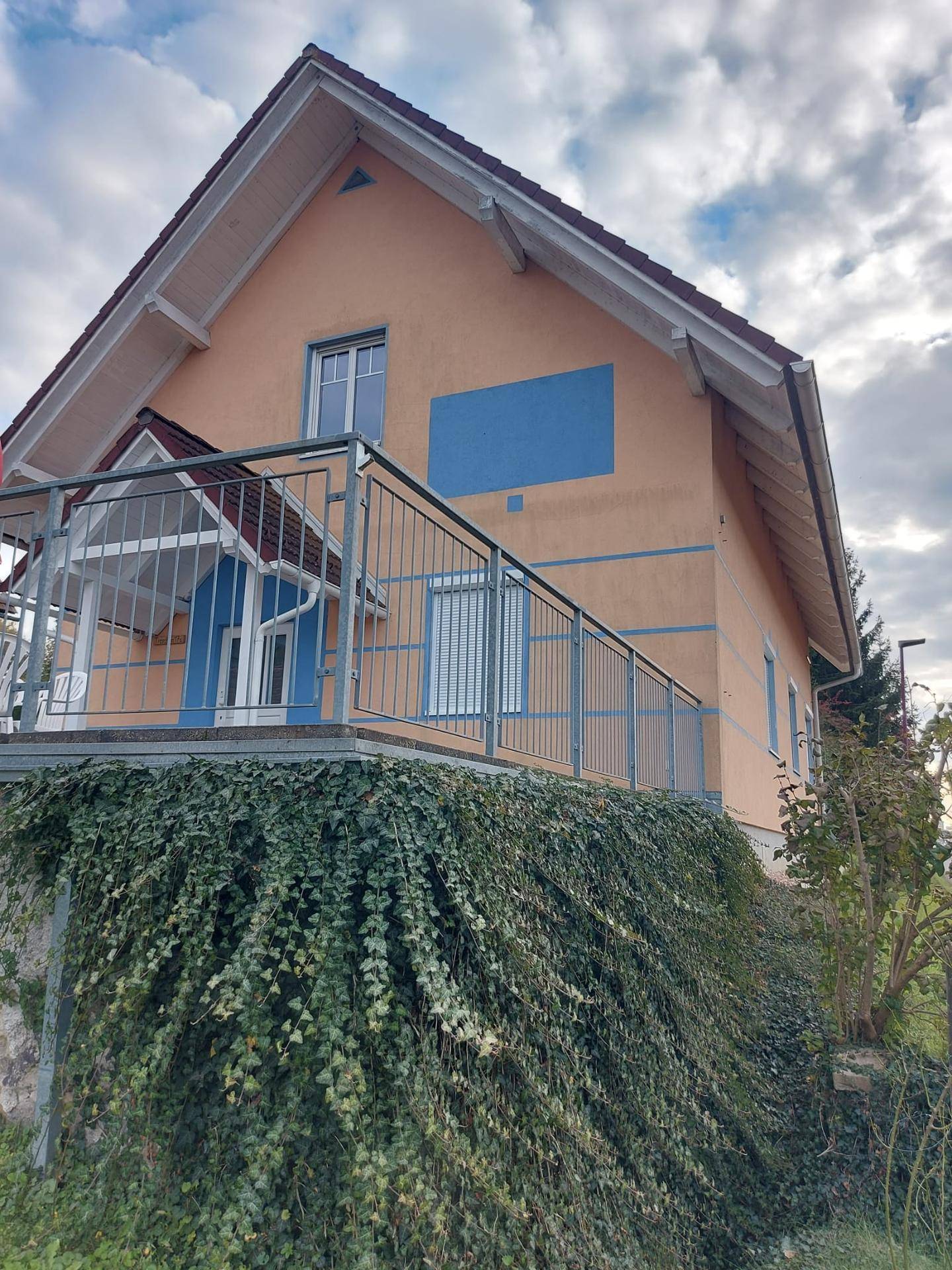 Seitenansicht des Hauses mit erhöhter Terrasse und üppigem Efeu, das die Stützmauer bedeckt.