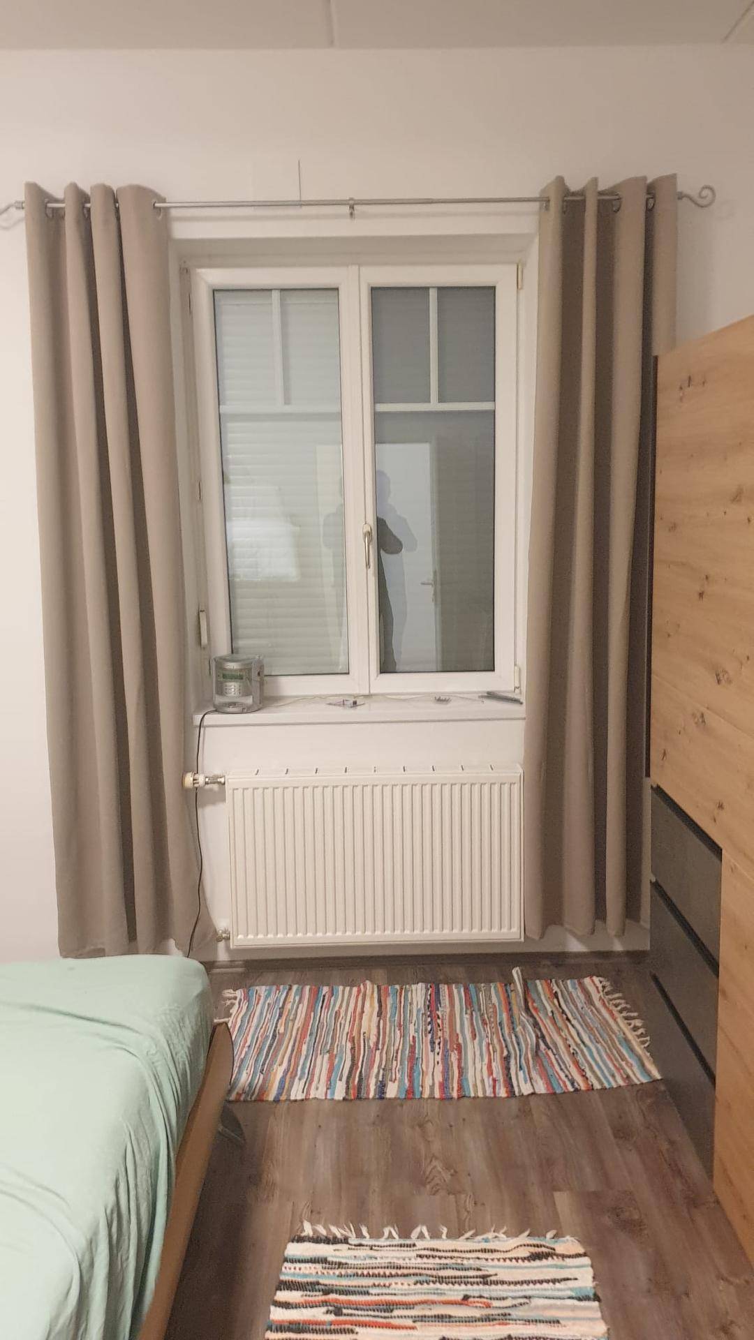 Fenster im Schlafzimmer mit beigen Vorhängen und Heizkörper, das viel Tageslicht hereinlässt.