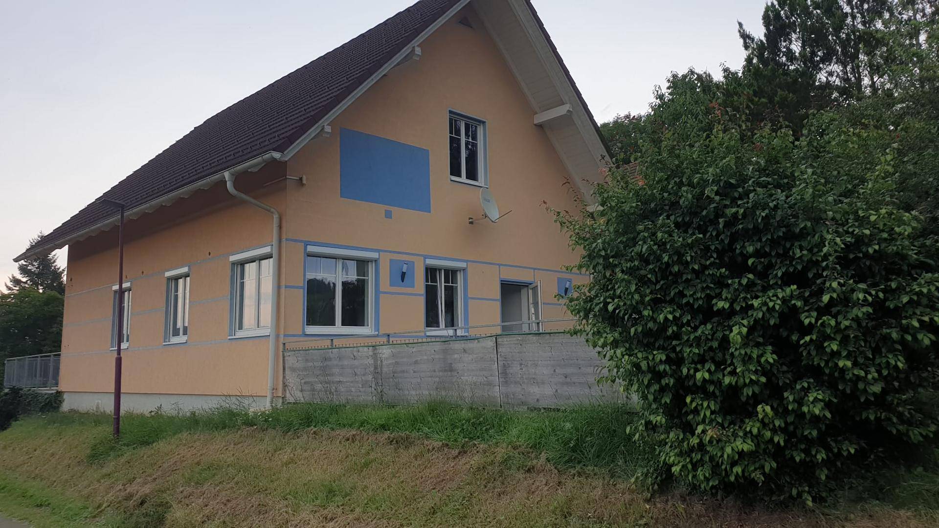 Ansicht des Hauses mit beiger Fassade und blauen Akzenten, umgeben von Bäumen und Grünflächen.