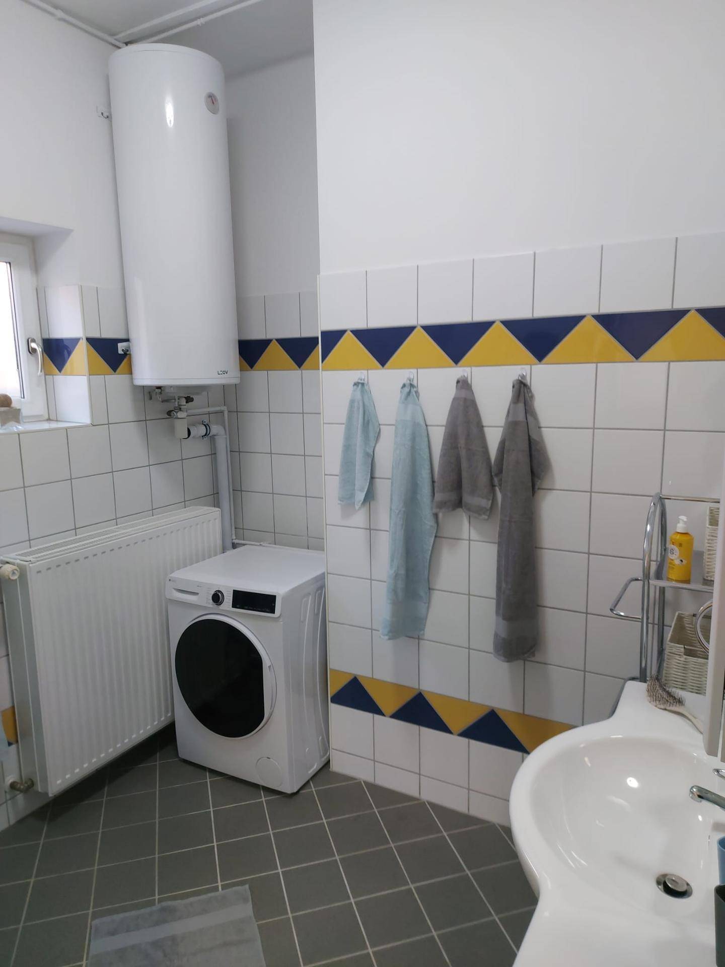 Badezimmer mit Waschmaschine, Warmwasserboiler und farbenfrohen Fliesen, sowie einem Fenster.