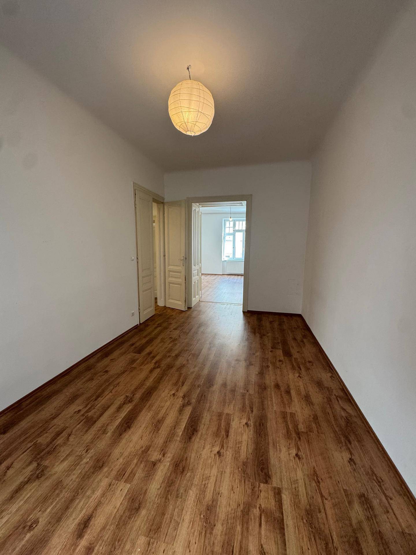 Leeres Zimmer mit Laminatboden und weiß gestrichenen Wänden, ein Durchgang führt in einen weiteren Raum.