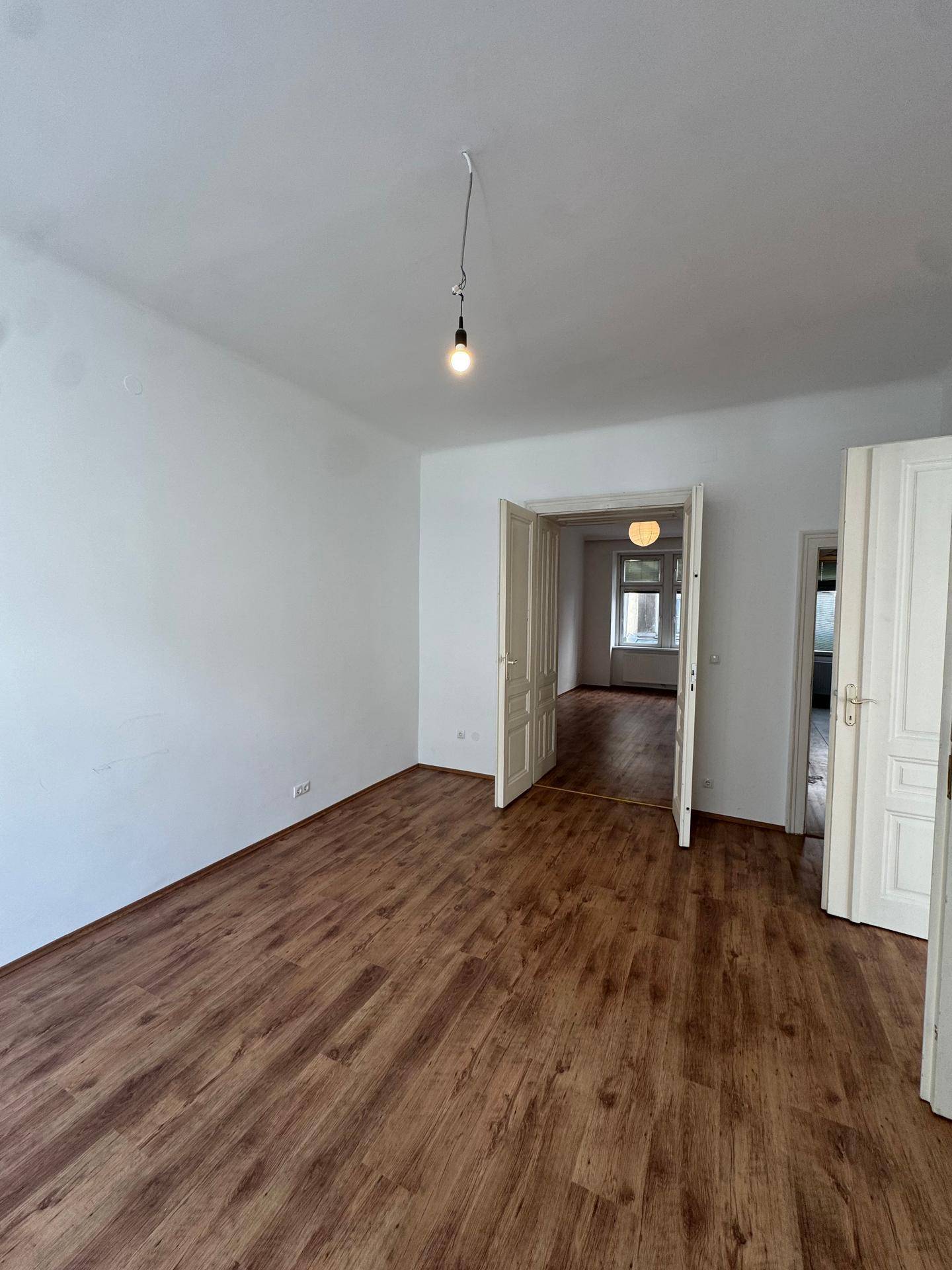 Helles, leeres Zimmer mit Laminatboden und weißen Wänden, das durch offene Türen mit anderen Räumen verbunden ist.