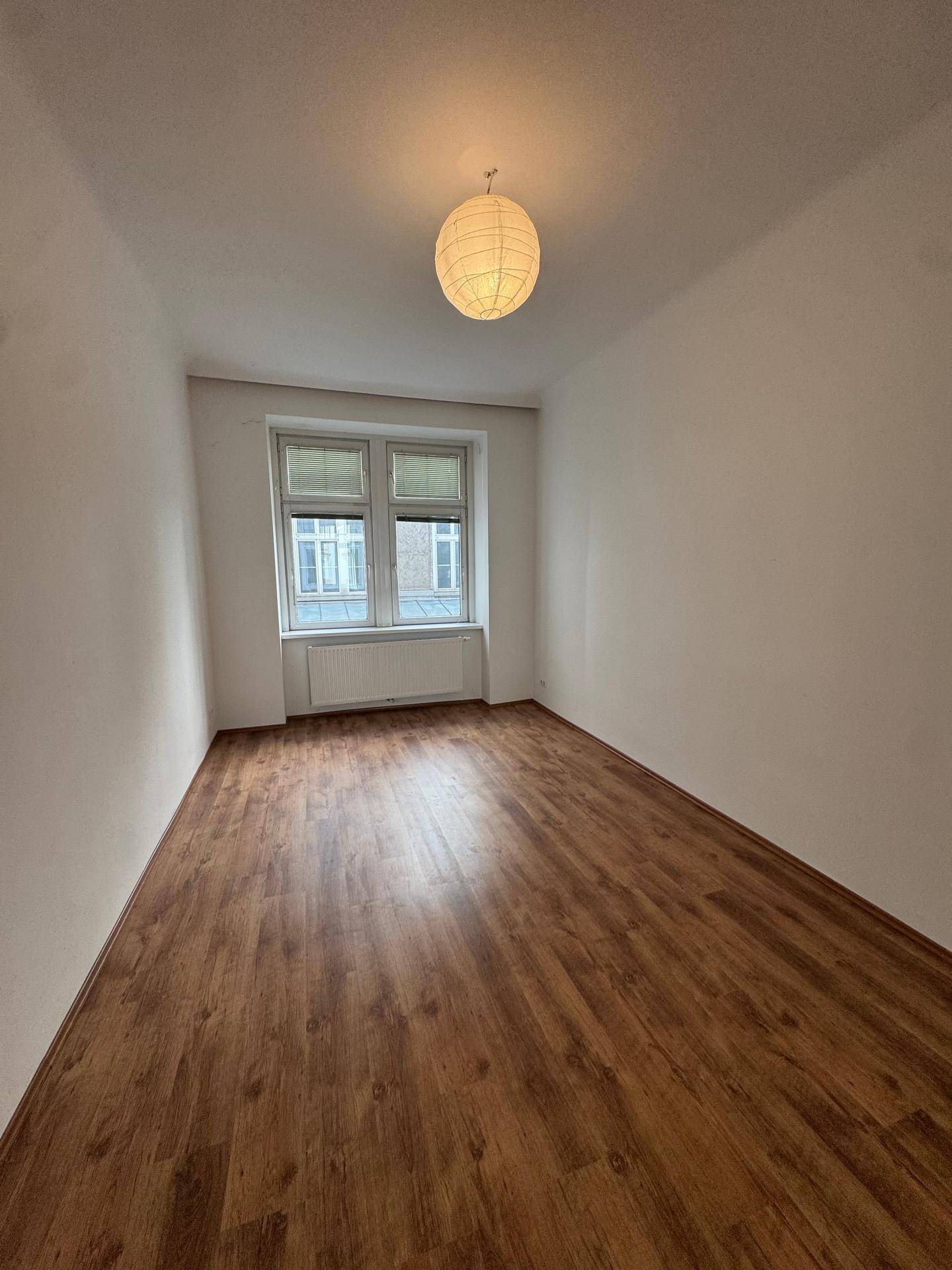 Helles, leeres Zimmer mit Laminatboden, weißen Wänden und einem Fenster, das Tageslicht hereinlässt.