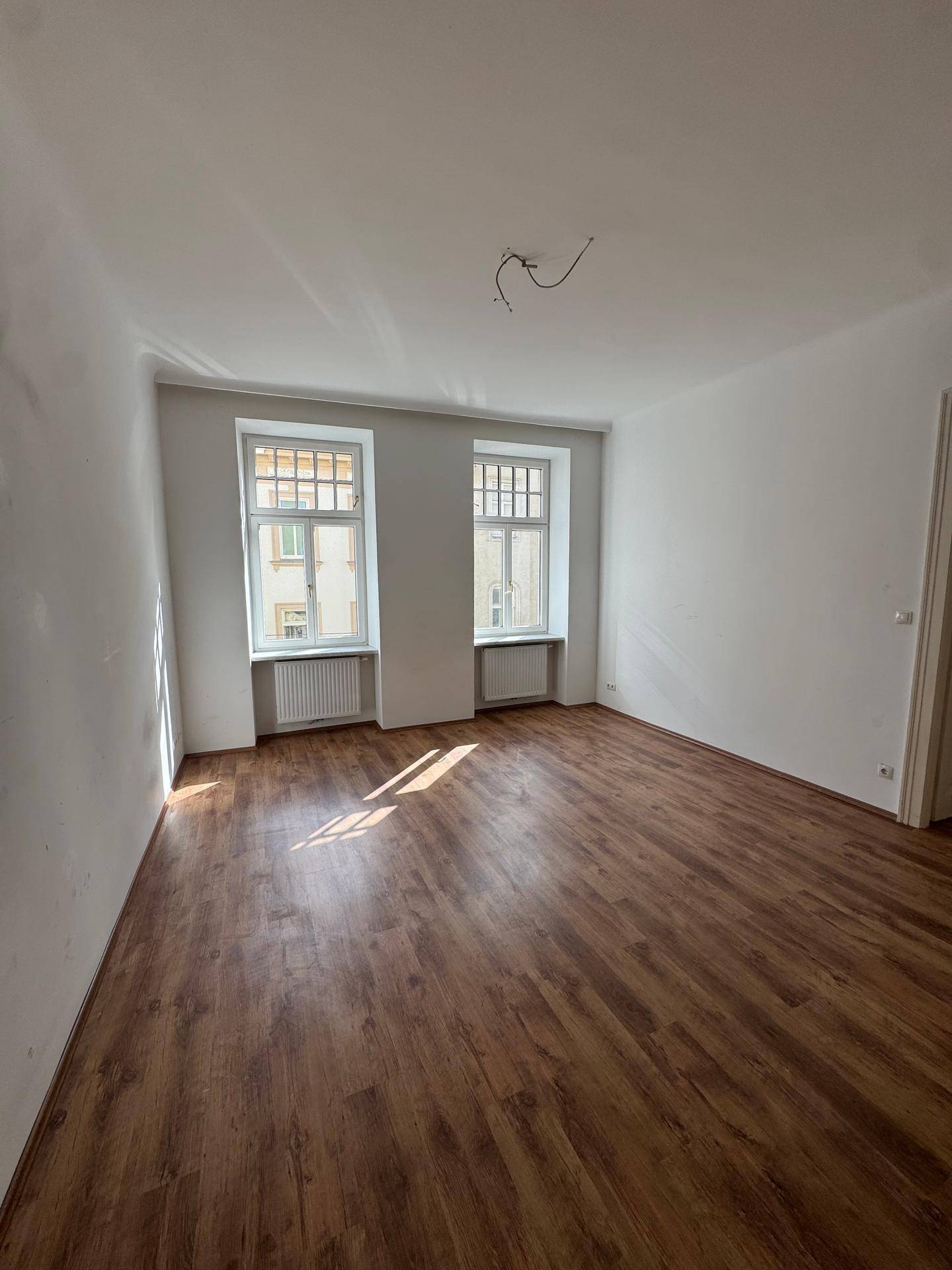 Geräumiges, leeres Zimmer mit Laminatboden, zwei großen Fenstern und viel natürlichem Lichteinfall.