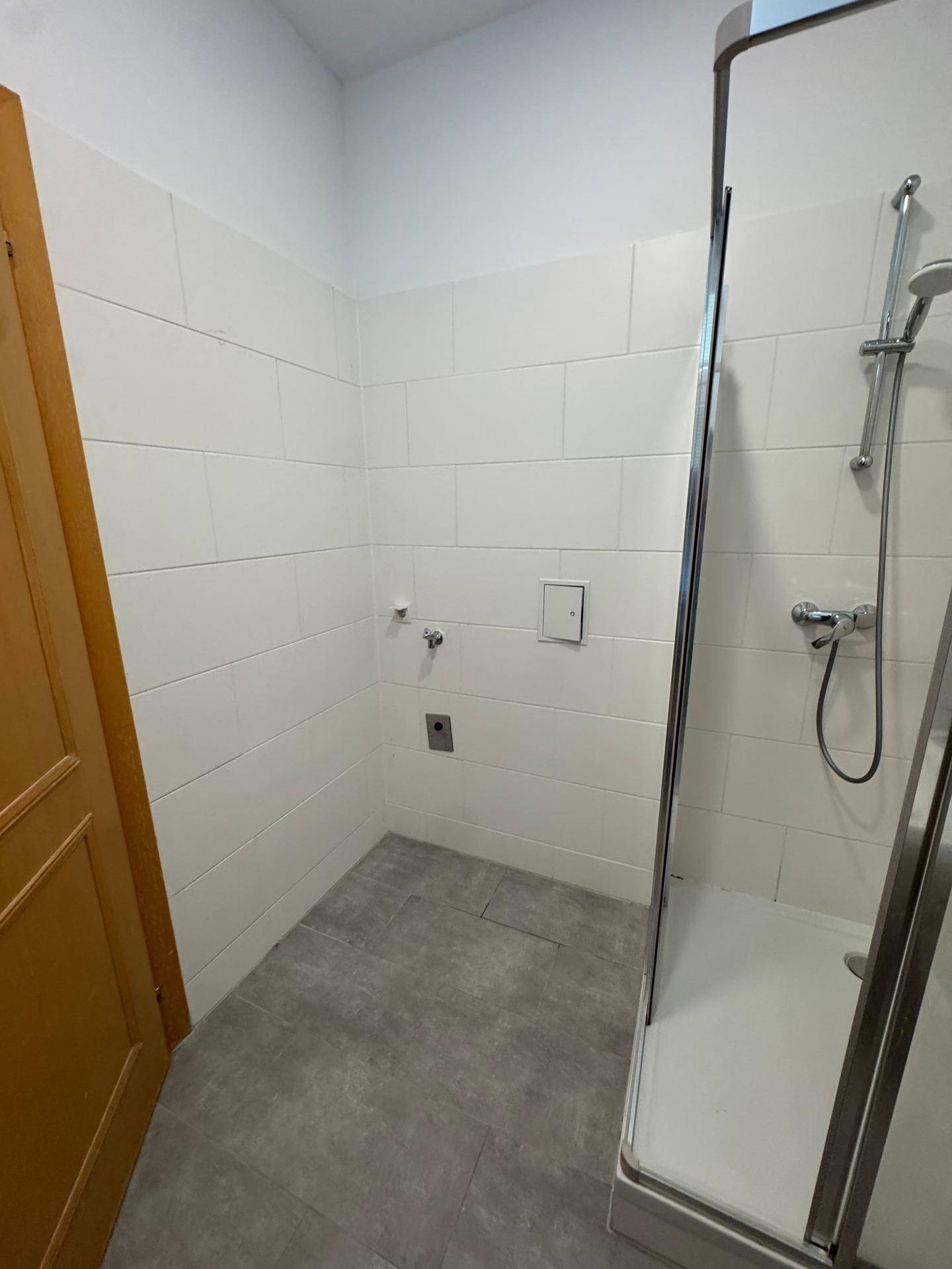 Badezimmer mit grauen Fliesen und weißen Wänden, ausgestattet mit einer Duschkabine und Anschlüssen.