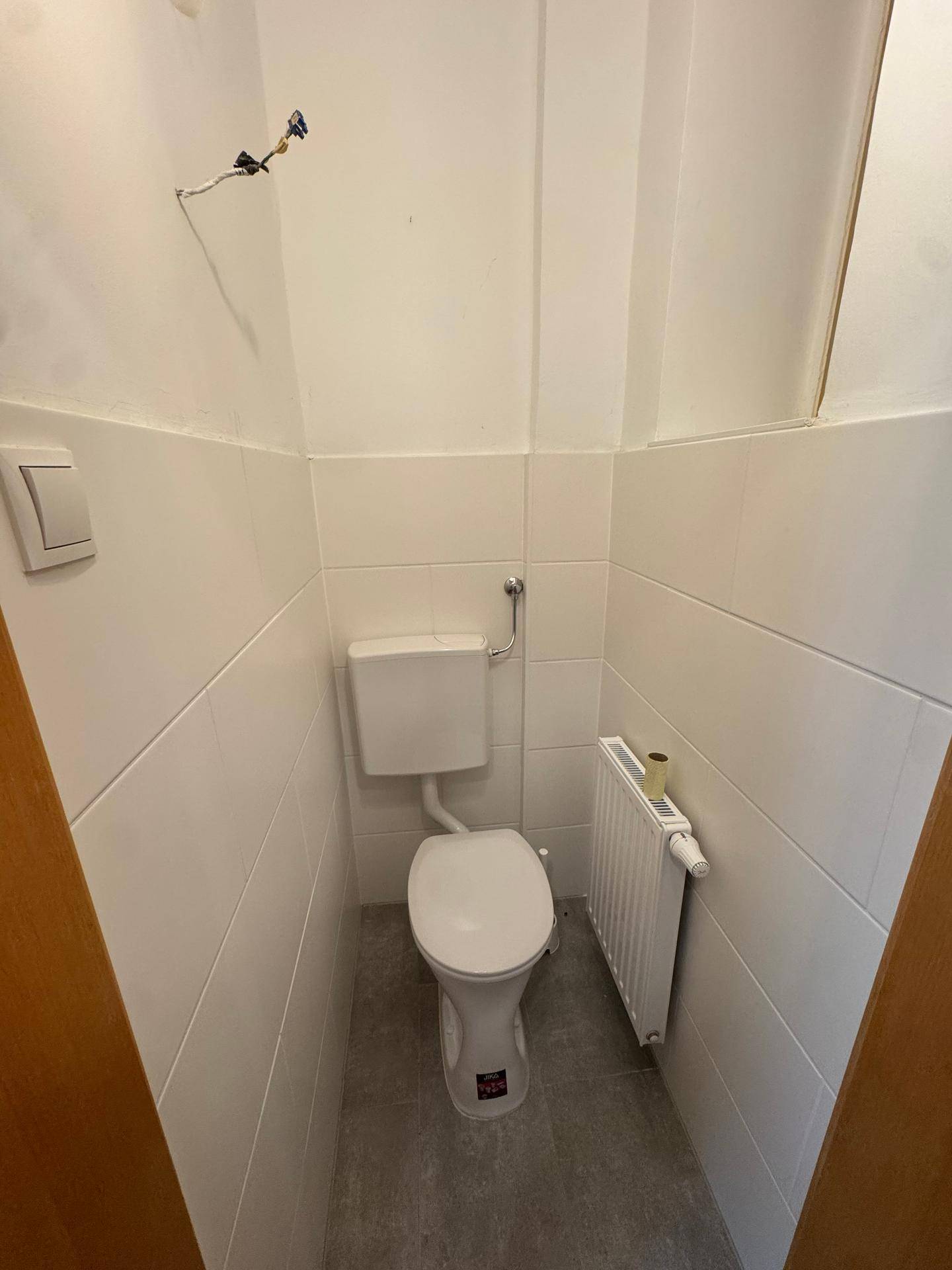 Kleines WC mit grauen Fliesen, weißen Wänden und einer Toilette, ausgestattet mit einem Heizkörper.