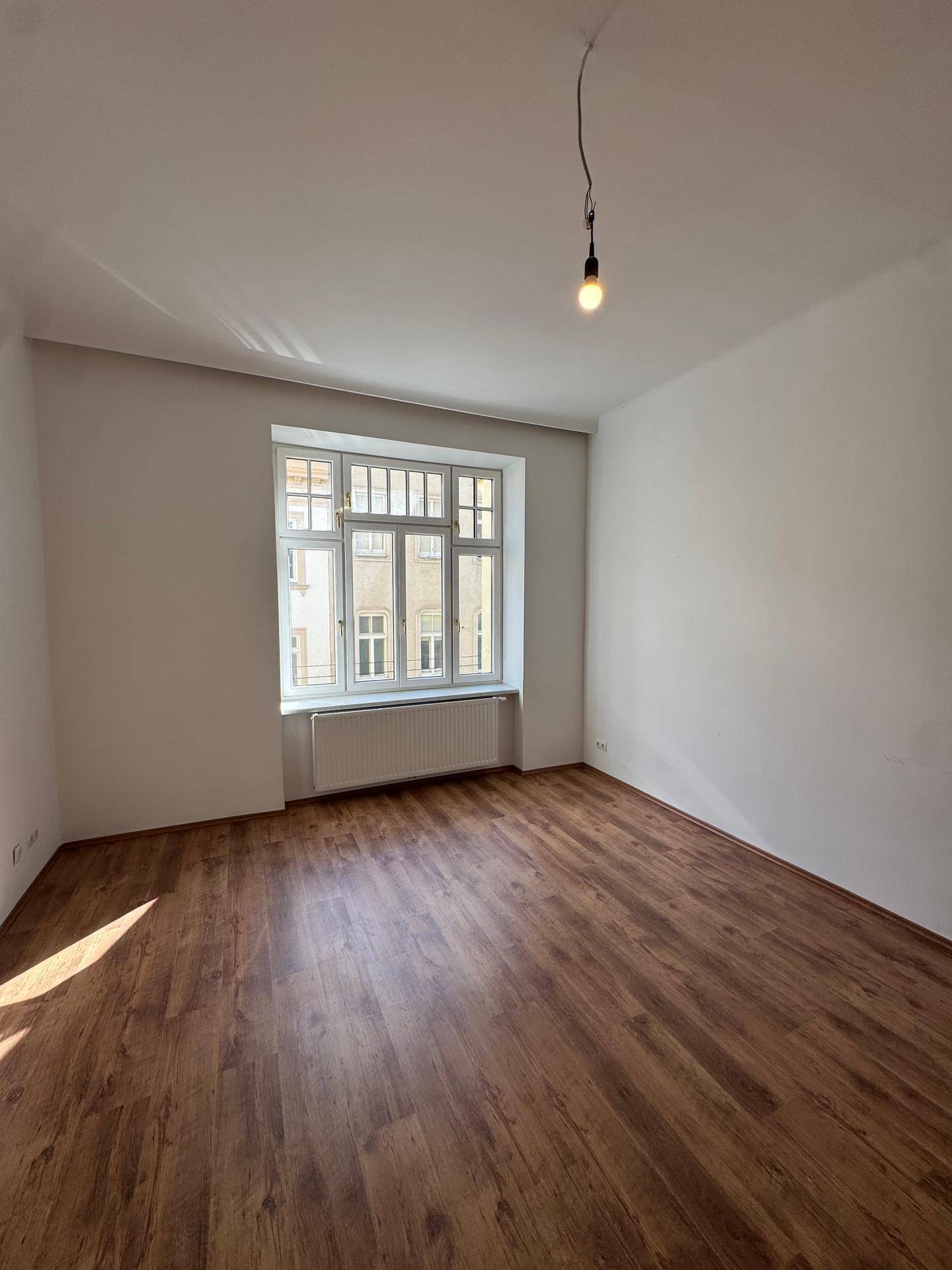 Sonniger, leerer Raum mit Laminatboden und einem großen Fenster, das viel natürliches Licht hereinlässt.
