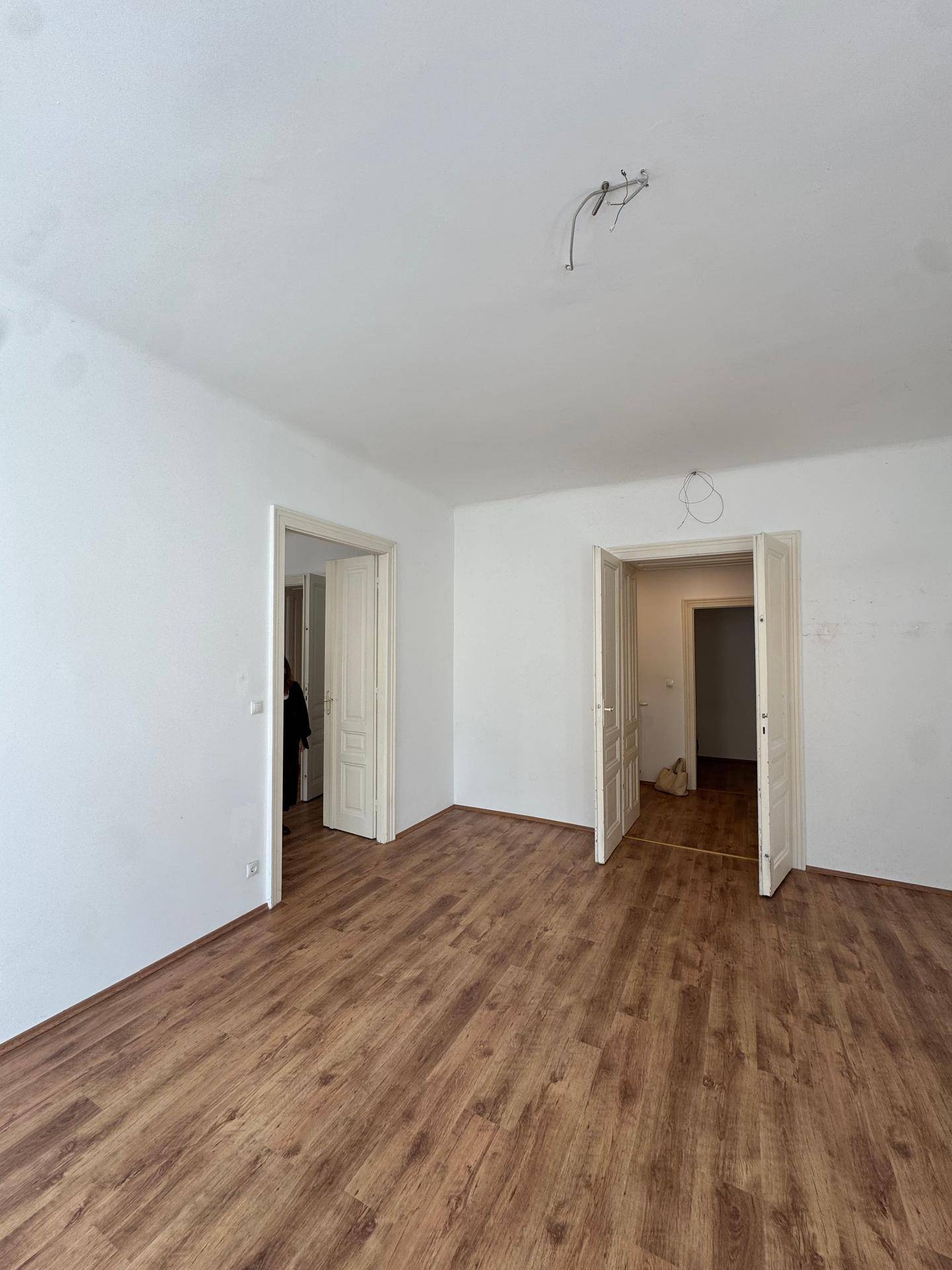 Geräumiges, leeres Zimmer mit Laminatboden und weißen Wänden, das durch mehrere Türen zugänglich ist.