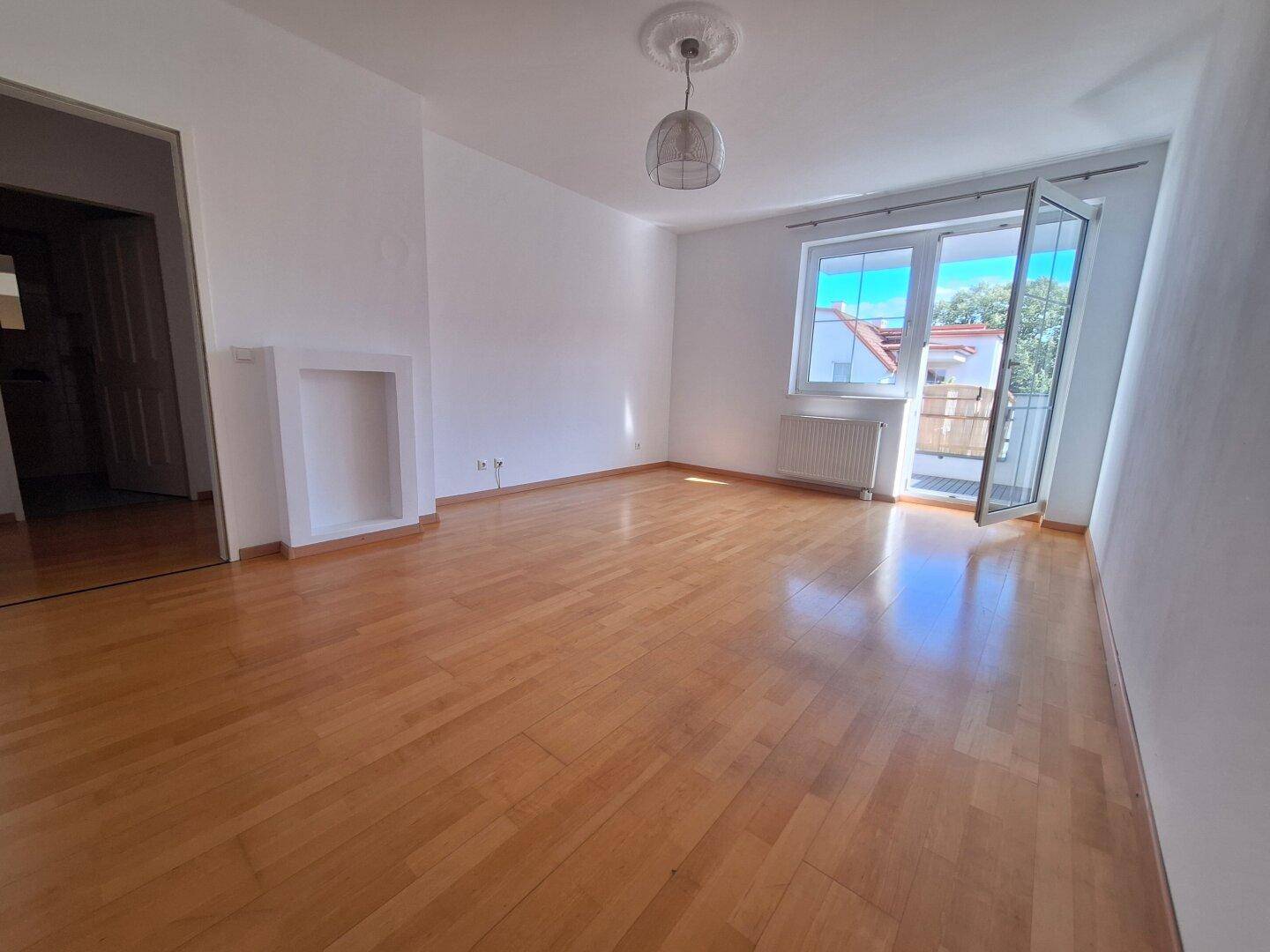 Helles Wohnzimmer mit Parkettboden, weißen Wänden und Zugang zu einem Balkon.