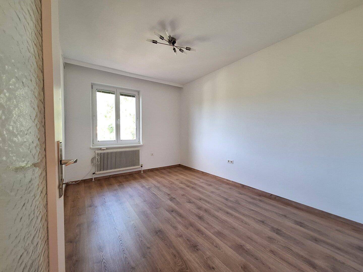 Leeres Schlafzimmer mit Laminatboden, weißen Wänden und einem großen Fenster.