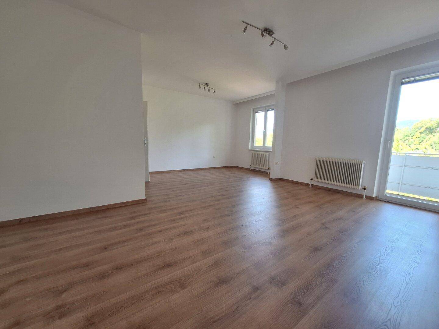 Helles, leeres Wohnzimmer mit Laminatboden, weißen Wänden und Zugang zum Balkon.
