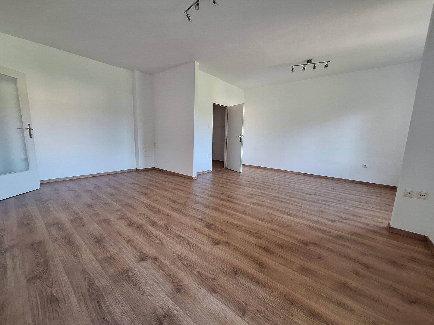 Geräumiges Wohnzimmer mit hellem Laminatboden und frisch gestrichenen weißen Wänden.