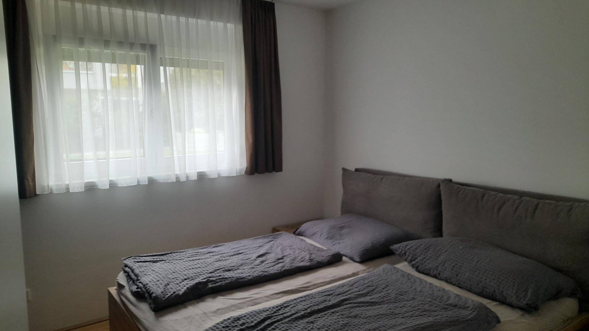 Schlafzimmer mit Doppelbett, grauer Bettwäsche und einem Fenster mit Vorhängen.