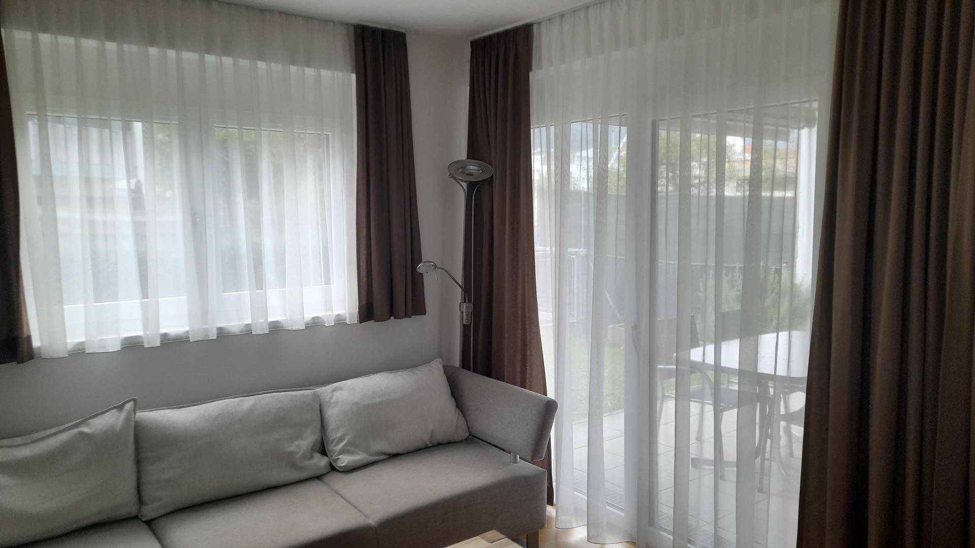 Helles Wohnzimmer mit grauem Sofa, Vorhängen und Zugang zu einem Außenbereich.