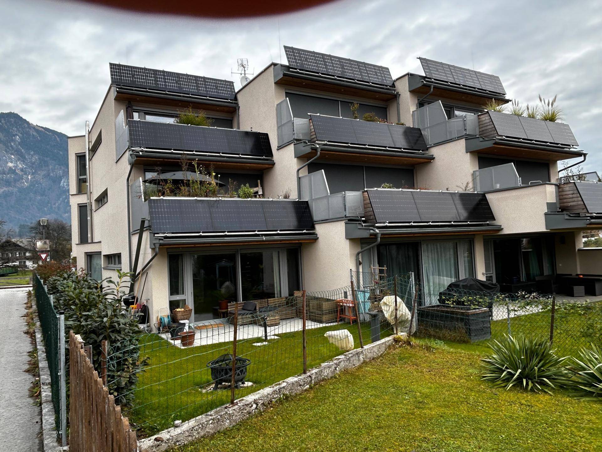 Modernes Mehrfamilienhaus mit Photovoltaikanlagen, großzügigen Balkonen und Terrassen, umgeben von Grünflächen und mit Bergblick.