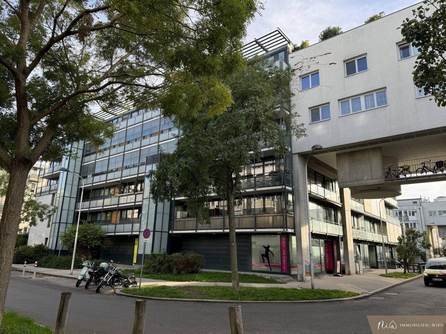 Ansicht eines modernen Wohngebäudes mit Glasfassade und Bäumen an der Straße.