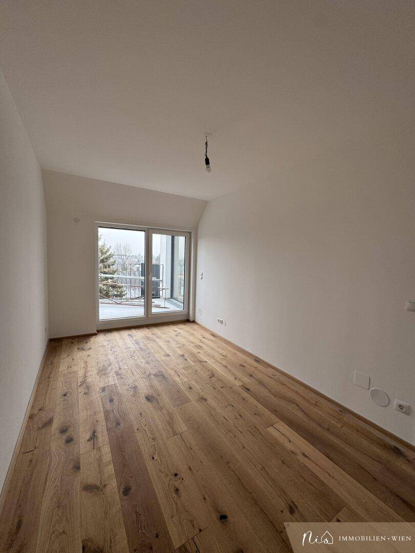 Helles, leeres Zimmer mit Holzboden und Fenster mit Blick nach draußen.