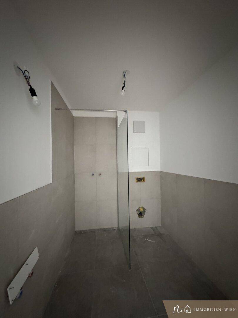Unfertiger Badezimmerbereich mit grauen Fliesen und einer Glaswand für die Dusche.