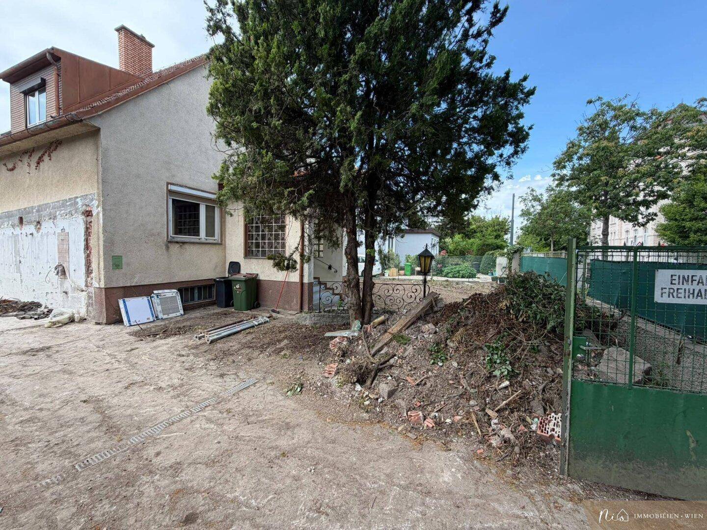 Seitenansicht des Hauses mit einem Baum, Bauschutt und einem grünen Gartentor.