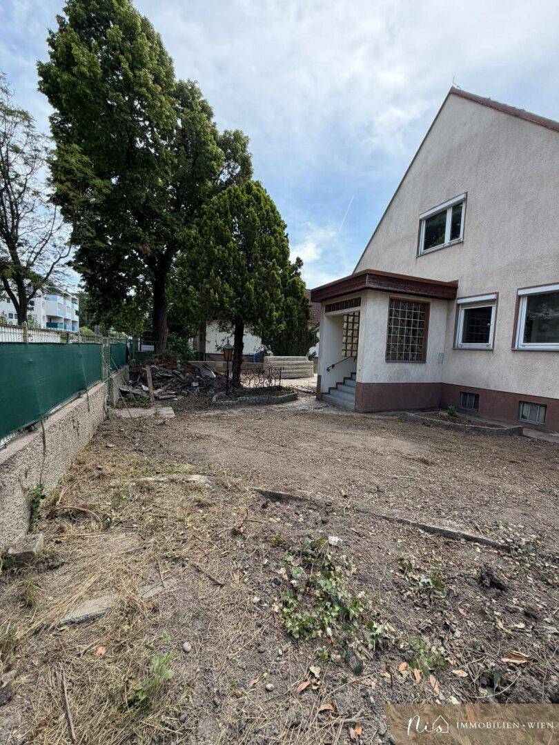 Seitenansicht des Hauses mit einem überdachten Eingang und hohen Bäumen im Garten.