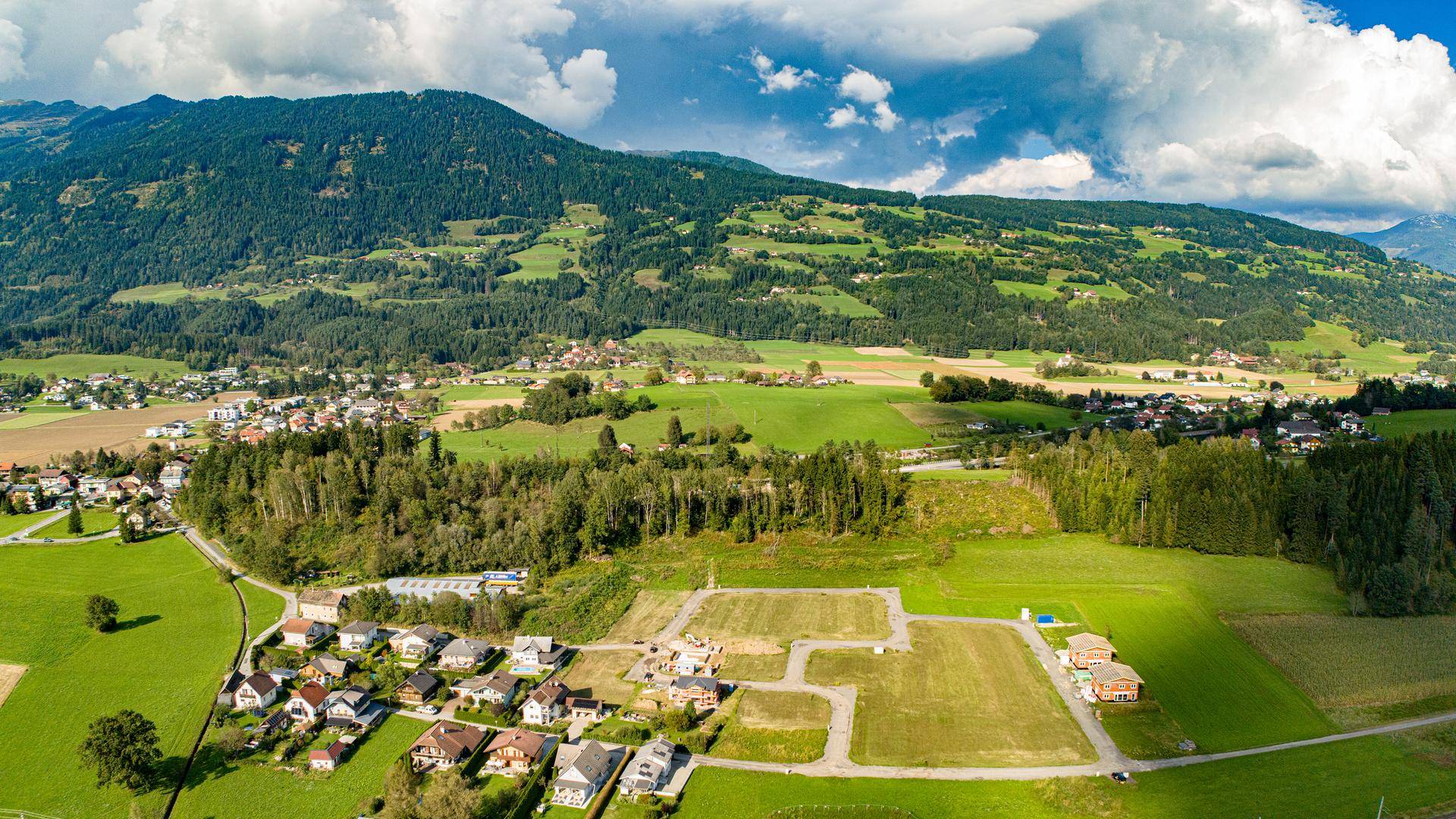 Weitwinkel-Luftaufnahme einer Wohnsiedlung in einer malerischen Berglandschaft mit Feldern und Wäldern.