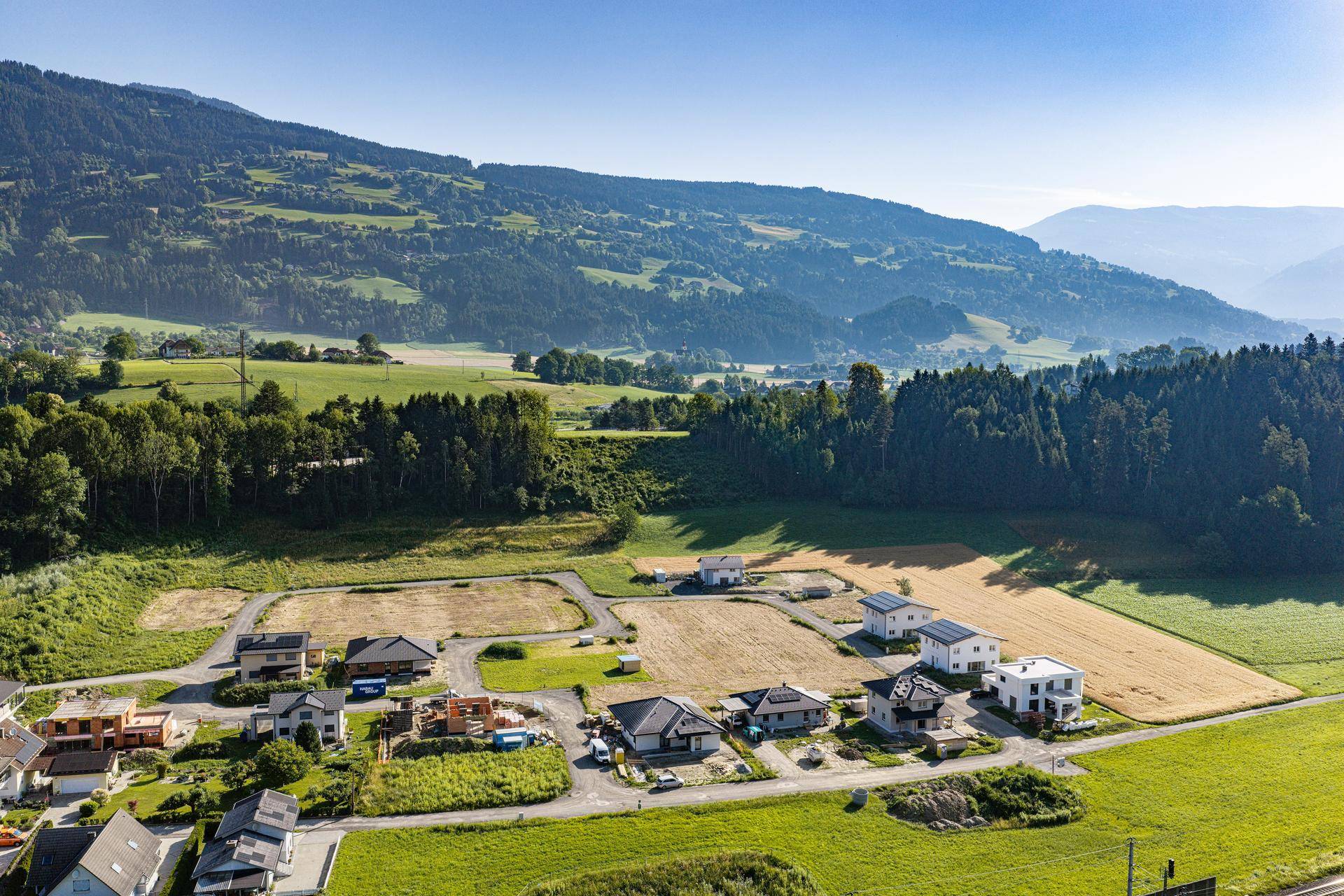 Weitläufige Luftaufnahme eines Wohngebietes in Entwicklung, umgeben von grünen Feldern und bewaldeten Bergen.
