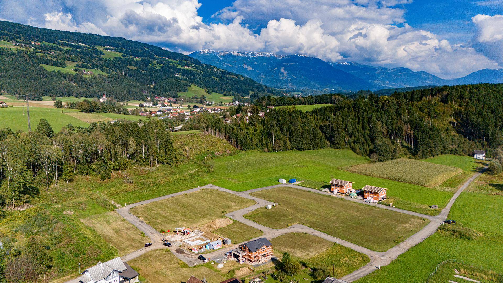 Panoramablick auf ein Neubaugebiet mit Bauprojekten und freien Grundstücken, umgeben von grünen Hügeln und schneebedeckten Bergen.