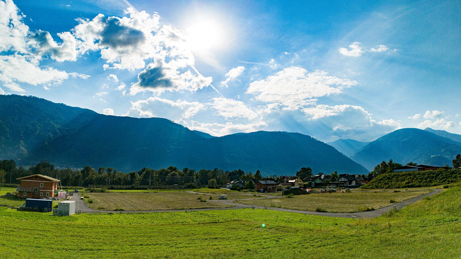 Sonniger Panoramablick auf ein Baugebiet mit grünen Feldern und beeindruckender Bergkulisse.