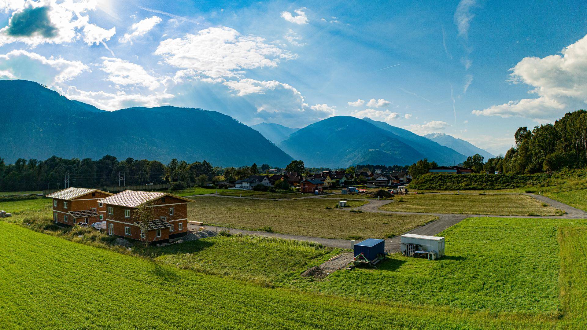 Sonniger Panoramablick auf ein ländliches Baugebiet mit zwei neuen Häusern und majestätischen Bergen im Hintergrund.