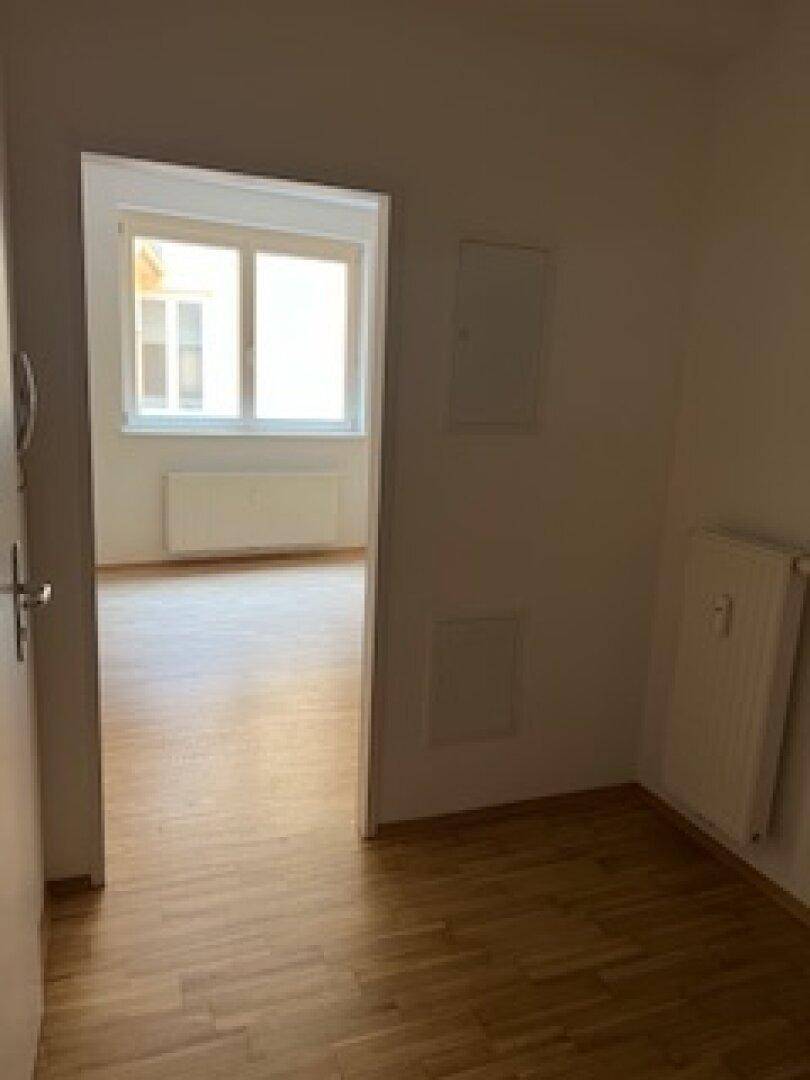 Blick aus einem Flur in einen hellen, leeren Raum mit Fenster und durchgehendem Holzfußboden.