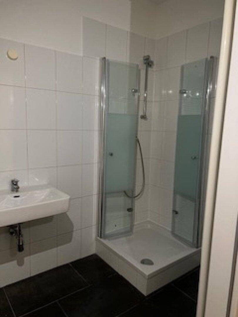 Funktionales Badezimmer mit Duschkabine, Waschbecken und weißen Wandfliesen auf dunklem Fliesenboden.