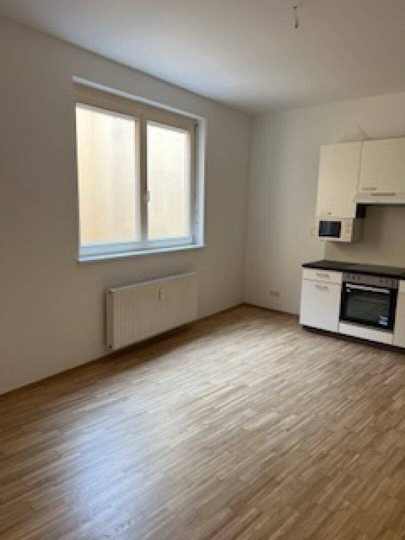 Heller Wohnraum mit großem Fenster, Heizkörper und Holzfußboden, teilweise mit Küchenzeile.