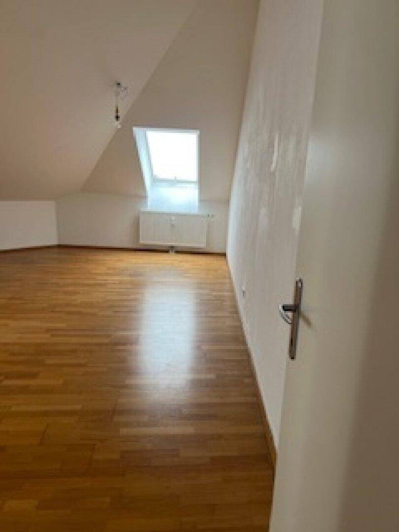 Geräumiger Dachgeschossraum mit Parkettboden, weiß gestrichenen Wänden und einem Dachfenster.