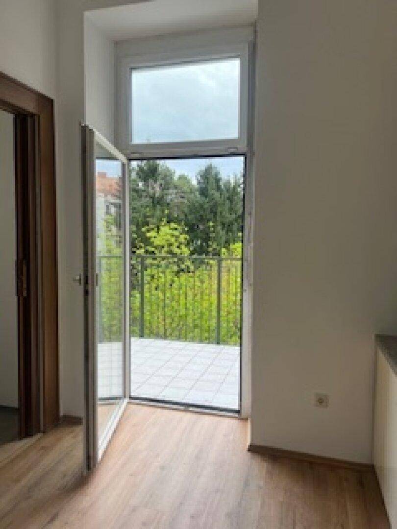 Heller Raum mit Holzboden und Zugang zu einem Balkon mit Blick ins Grüne.