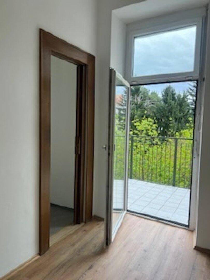 Leerer Raum mit Holzboden und Zugang zu einem Balkon mit grüner Aussicht.